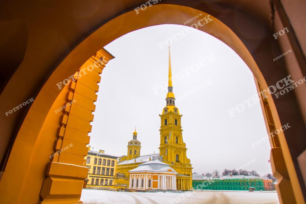 На фотографии изображен Петропавловский собор, расположенный на территории Петропавловской крепости в Санкт-Петербурге. Золотой шпиль собора выделяется на фоне зимнего неба, а рамка из арки крепости добавляет глубины композиции. Петропавловский собор — одно из главных архитектурных сооружений города, символ Санкт-Петербурга и место захоронения русских императоров. Зимний снег подчеркивает строгую красоту архитектуры.